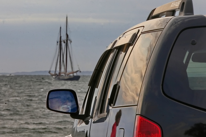 A man is seen reflected in his rear-view mirror looking at the schooner Ernestina-Morrissey as it heads into Buzzards Bay as a woman walking across Fort Phoenix in Fairhaven is reflected on the side of his vehicle. PHOTO PETER PEREIRA