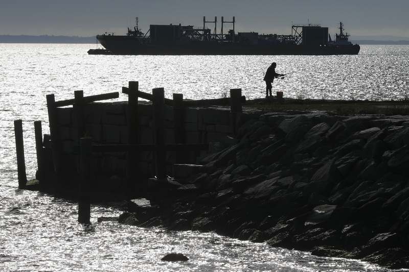 A man fishes from a pier on Fort Taber Park in New Bedford, MA as in the distance two tugboats maneuver a huge barge used by the offshore wind industry. PHOTO PETER PEREIRA