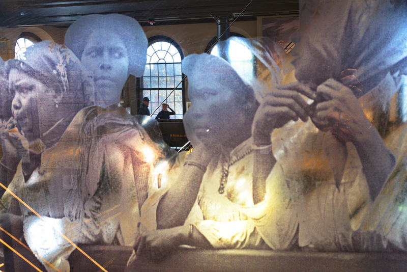 A transparency photo of Cape Verdean immigrants arriving in the United States in 1920 seen behind the model of the Ernestina-Morrissey schooner keep their eyes on a couple making their way across the upper level of the New Bedford Whaling Museum. PHOTO PETER PEREIRA