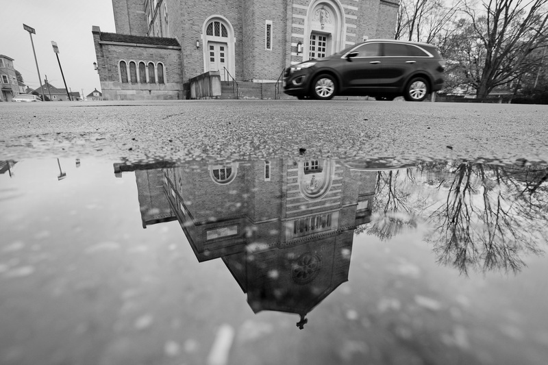 A vehicle moves down Acushnet Avenue as the reflection of the old St. Joseph & St. Therese Church appears in a puddle at the intersection of Acushnet Avenue and Deacon Street in New Bedford. The church is currently up for sale while the Diocese of Fall River keeps selling off properties due to a decline in parishioners. PHOTO PETER PEREIRA