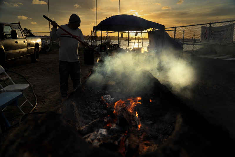 On October 21, 2026 the sun rises in the distance as Mashpee Wampanoag Tribe member Hartman Deetz tends the fire that hollows out a tree trunk, which will eventually become a mishoon canoe after several days of burning at the Community Boating Center in New Bedford, Massachusetts. PHOTO PETER PEREIRA